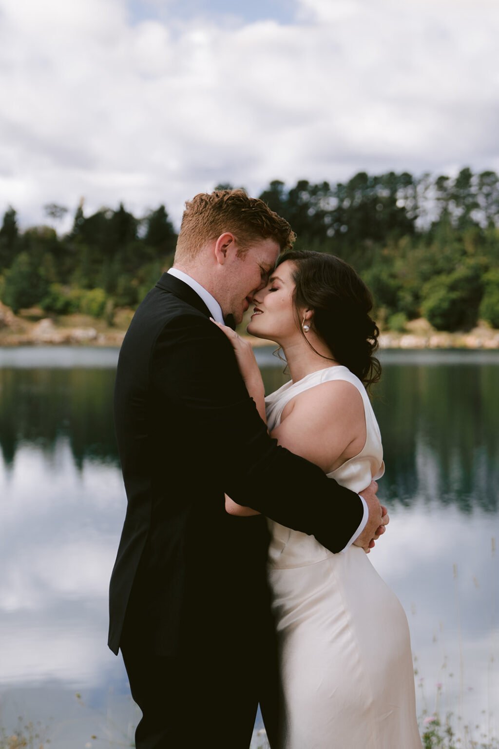 Bride and groom embracing by a lake in Portland, NSW, with the bride wearing minimal, natural makeup for a flawless look