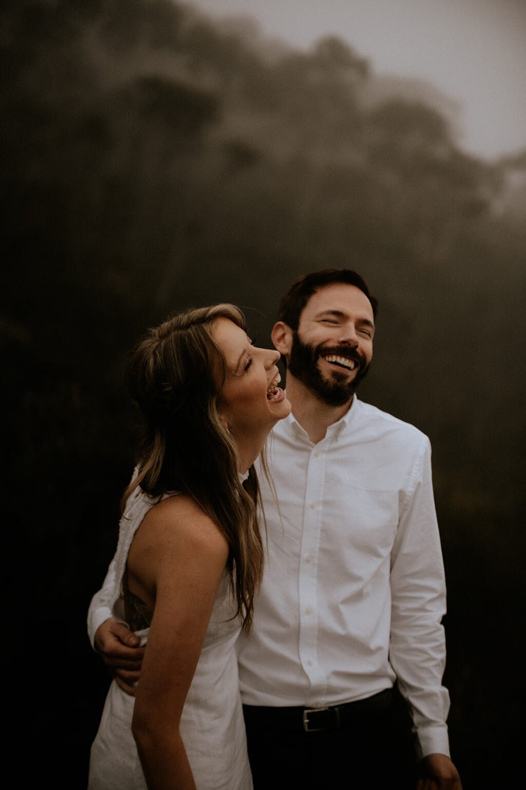 Happy couple outdoors, man in a white shirt and woman in a white dress, laughing together with their arms around each other.